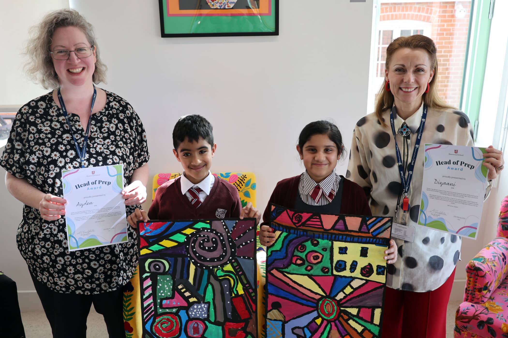 Two women and two pupils smile indoors; the women hold certificates, and the children, wearing school uniforms, proudly display colourful abstract artwork. Bright chairs and framed art are visible in the background.