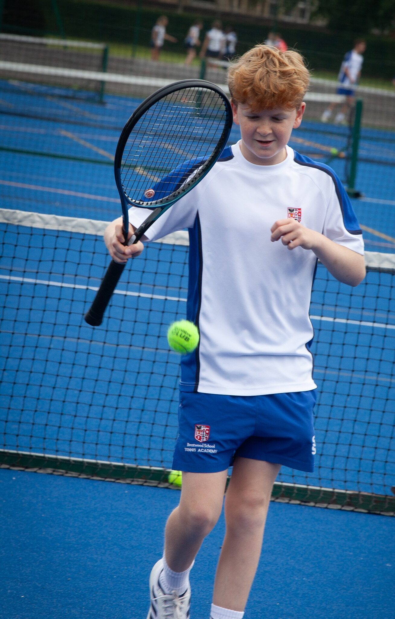 A young boy in a white and blue sports kit focuses on hitting a tennis ball with his racquet during a co-curricular match on a blue outdoor court. Other players are visible in the background.
