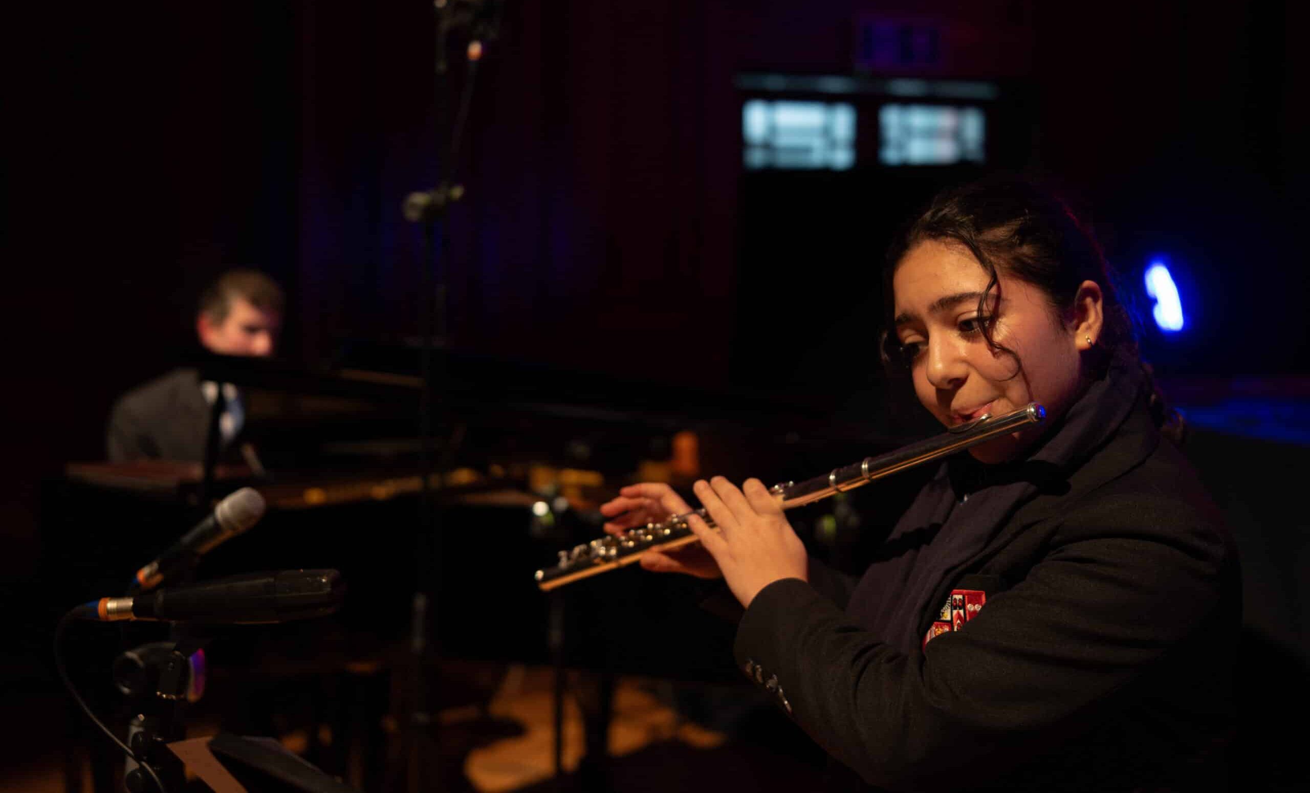 A young woman in a dark blazer plays the flute on stage, with microphones nearby, performing at music concert. In the background, a man sits at a piano in a dimly lit room, with a spotlight illuminating part of the scene.