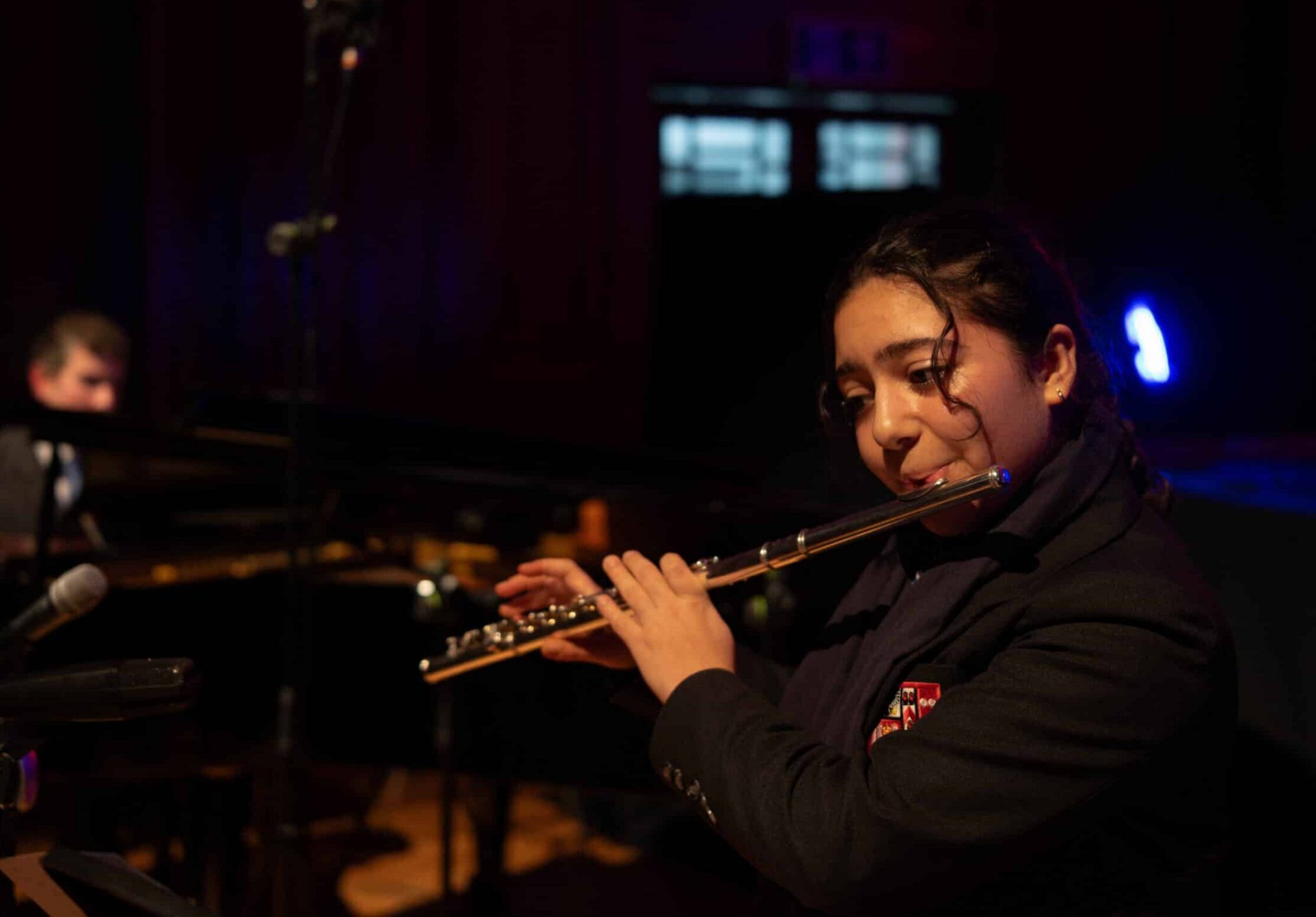 A young woman in a dark blazer plays the flute on stage, with microphones nearby, performing at music concert. In the background, a man sits at a piano in a dimly lit room, with a spotlight illuminating part of the scene.