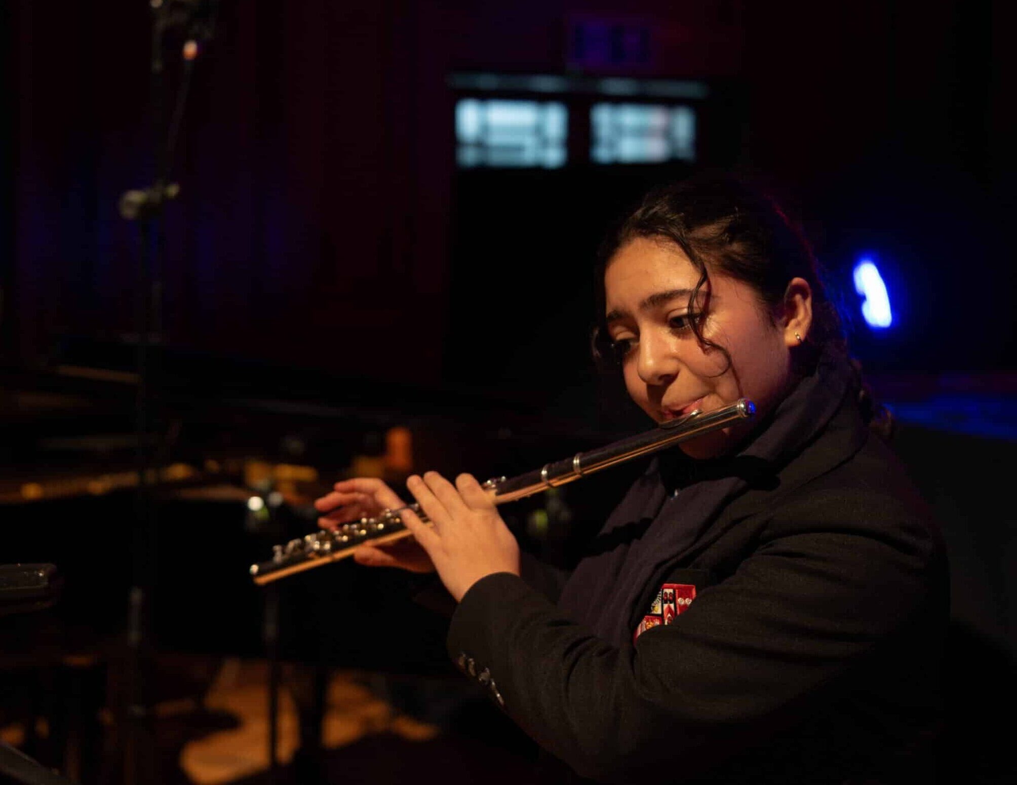 A young woman in a dark blazer plays the flute on stage, with microphones nearby, performing at music concert. In the background, a man sits at a piano in a dimly lit room, with a spotlight illuminating part of the scene.
