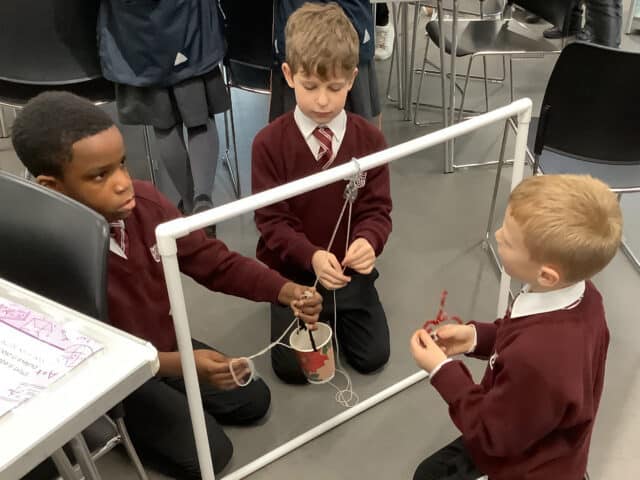 Three young boys in school uniforms kneel around a white frame with a pulley, working together to lift a tin bucket using string. Classroom chairs and a table with papers are visible in the background.