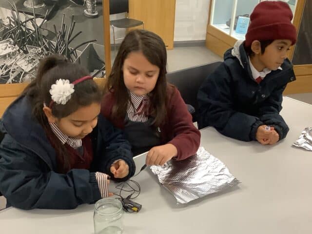 Three young children in school uniforms sit at a table working with sheets of aluminium foil and a battery pack. Two girls are focused on the foil, while a boy in a red hat sits beside them holding a pen.