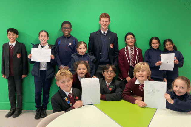 A group of eleven schoolchildren in uniforms stand and sit in front of a green wall. Four children seated at a table, and four standing behind them, hold up sketches on white paper and all are smiling at the camera.