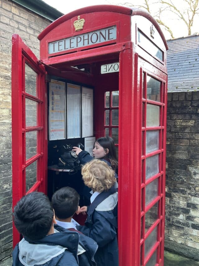 Three children stand outside a classic red British telephone box, watching a girl inside as she uses the black telephone. The box is open, and the children appear curious and engaged.