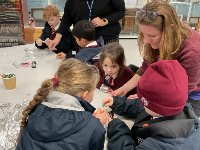 A group of young children, supervised by two adults, work together on a science project at a table. The children are wearing school uniforms and appear focused on assembling materials.