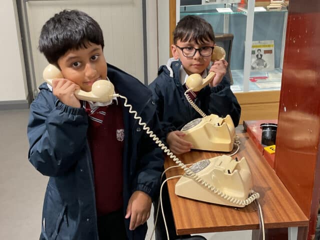 Two boys in school uniforms use vintage rotary dial telephones at a wooden table in what appears to be a museum or classroom setting. They are both holding the receivers to their ears.