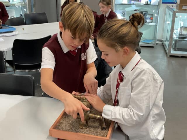 Two schoolchildren in uniform work together on a practical science activity with metal pipes at a table, whilst other pupils in similar uniforms are seen in the background.