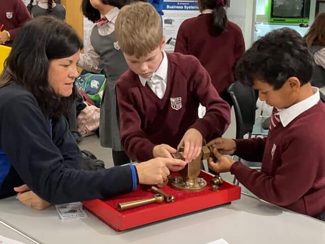 A woman assists two schoolchildren in maroon uniforms as they work together on a hands-on brass science experiment at a table in a classroom or workshop setting.