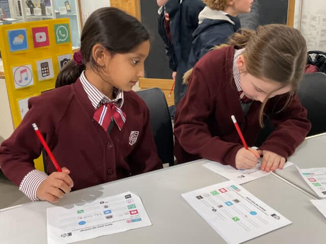 Two schoolgirls in uniforms sit at a table, working on worksheets with red pencils. One girl is writing whilst the other looks at her. In the background, a colourful display shows app icons.