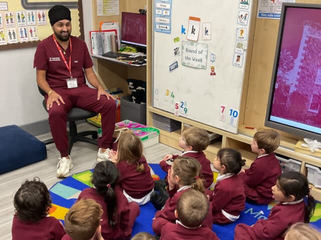 A man in maroon scrubs sits on a chair in front of a whiteboard, speaking to a group of young children in matching maroon uniforms seated on a colourful classroom rug.