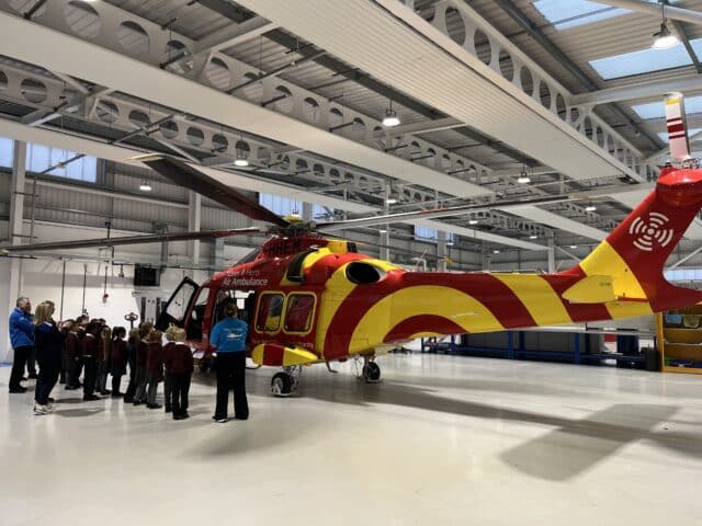 A group of children and adults stand next to a large yellow and red air ambulance helicopter inside a spacious hangar with high ceilings and bright lighting.