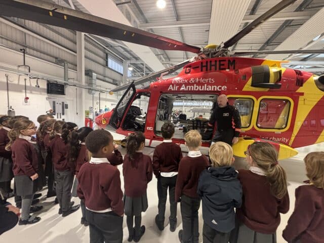 A group of schoolchildren in uniform listen to a person explaining next to a red and yellow Essex & Herts Air Ambulance helicopter inside a hangar.