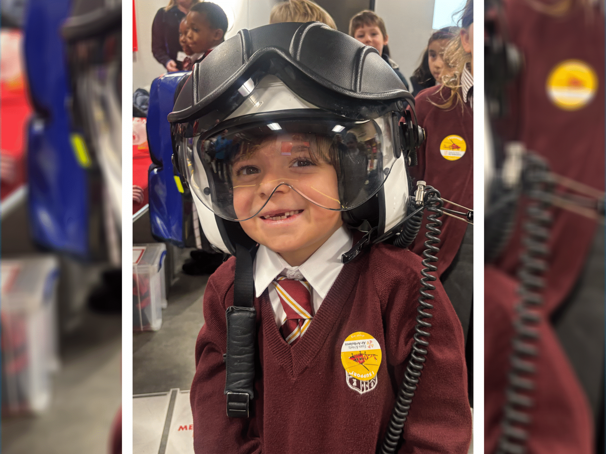 A young child in a maroon school uniform and striped tie smiles while wearing a large helicopter helmet with a visor. Other children in similar uniforms are visible in the background.