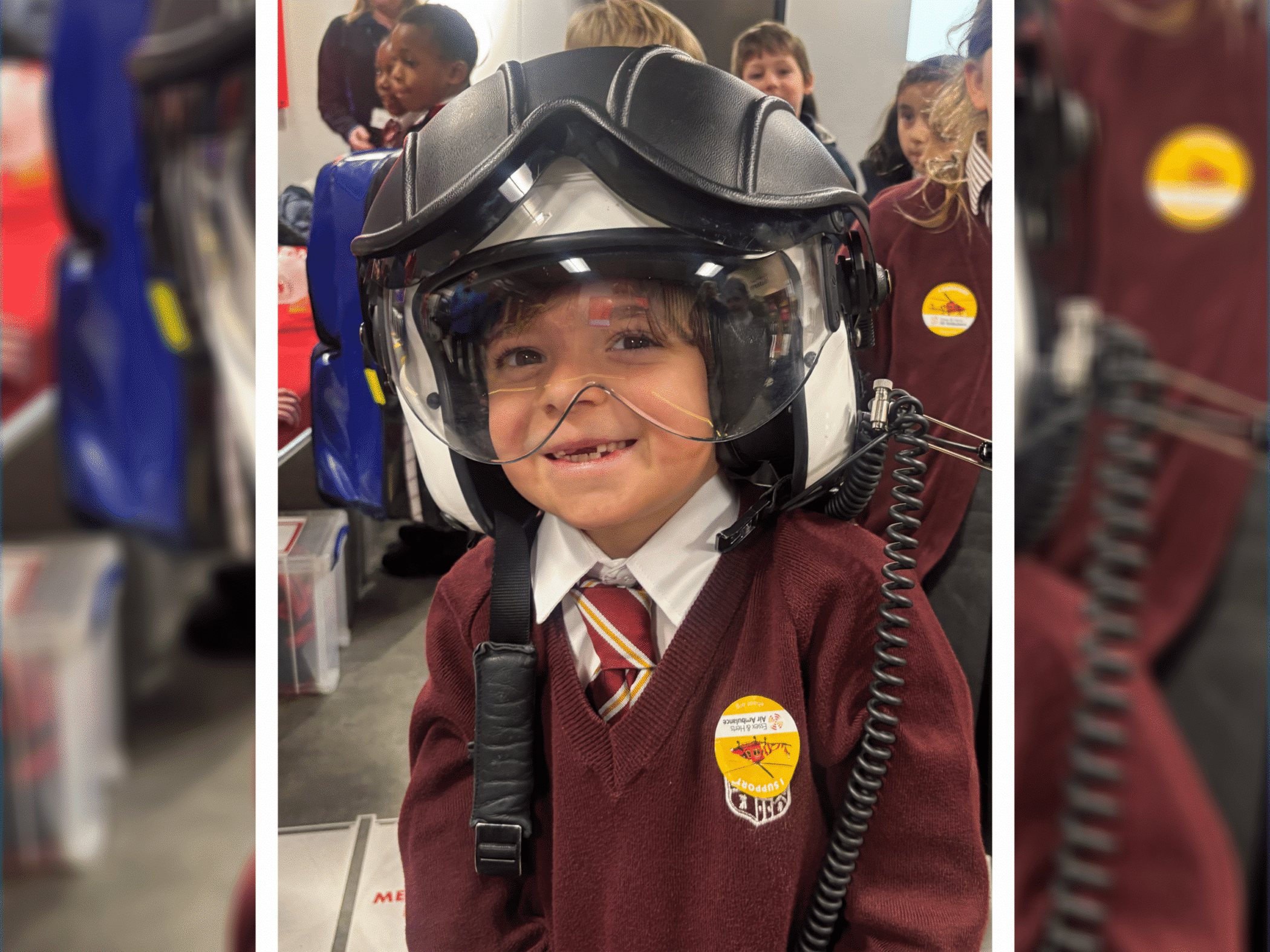 A young child in a maroon school uniform and striped tie smiles while wearing a large helicopter helmet with a visor. Other children in similar uniforms are visible in the background.