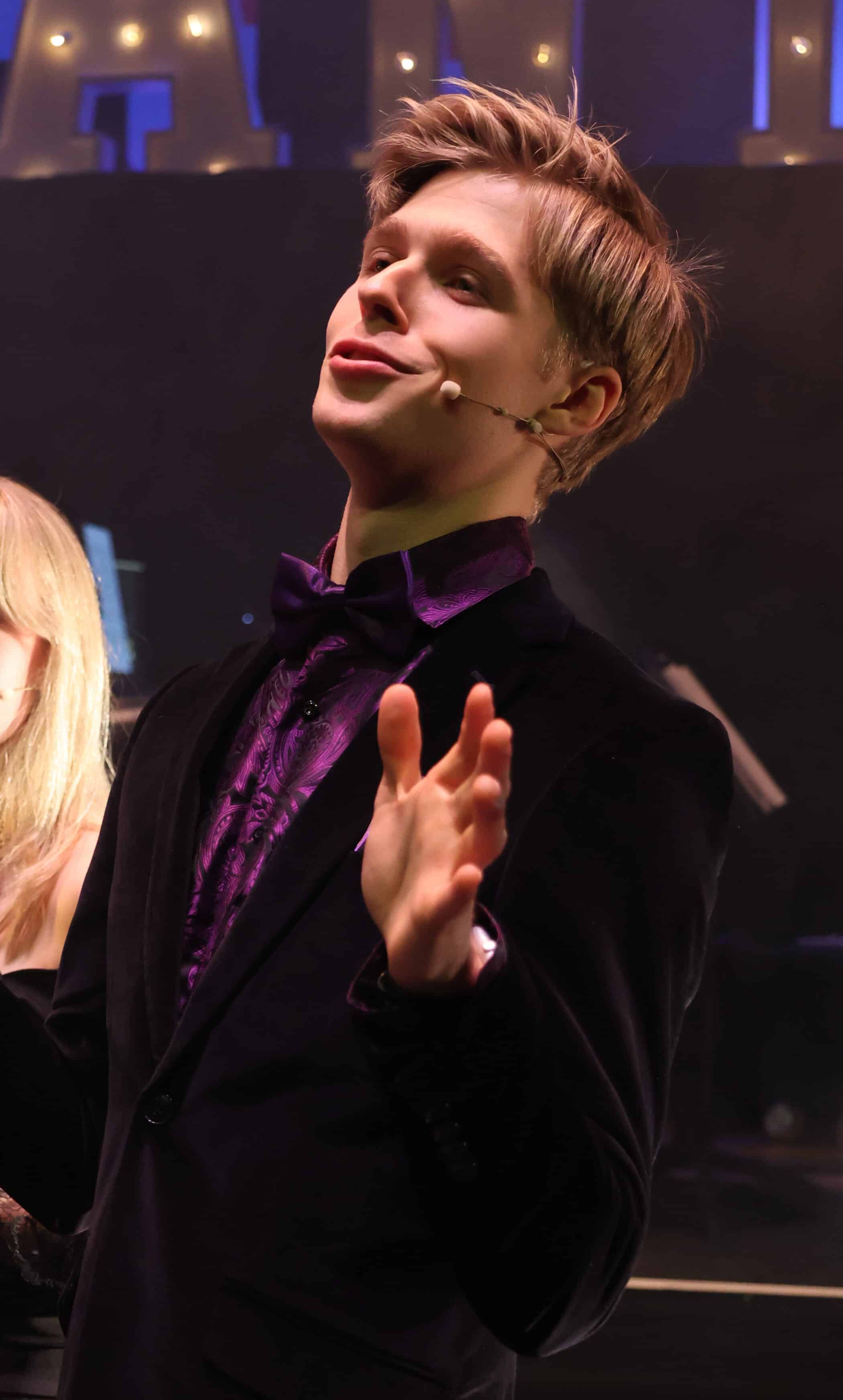 A young man with light brown hair wearing a dark velvet suit and a purple shirt speaks animatedly on stage about scholarships, with a headset microphone on his face and his hands raised mid-gesture. Stage lights and instruments are visible in the background.