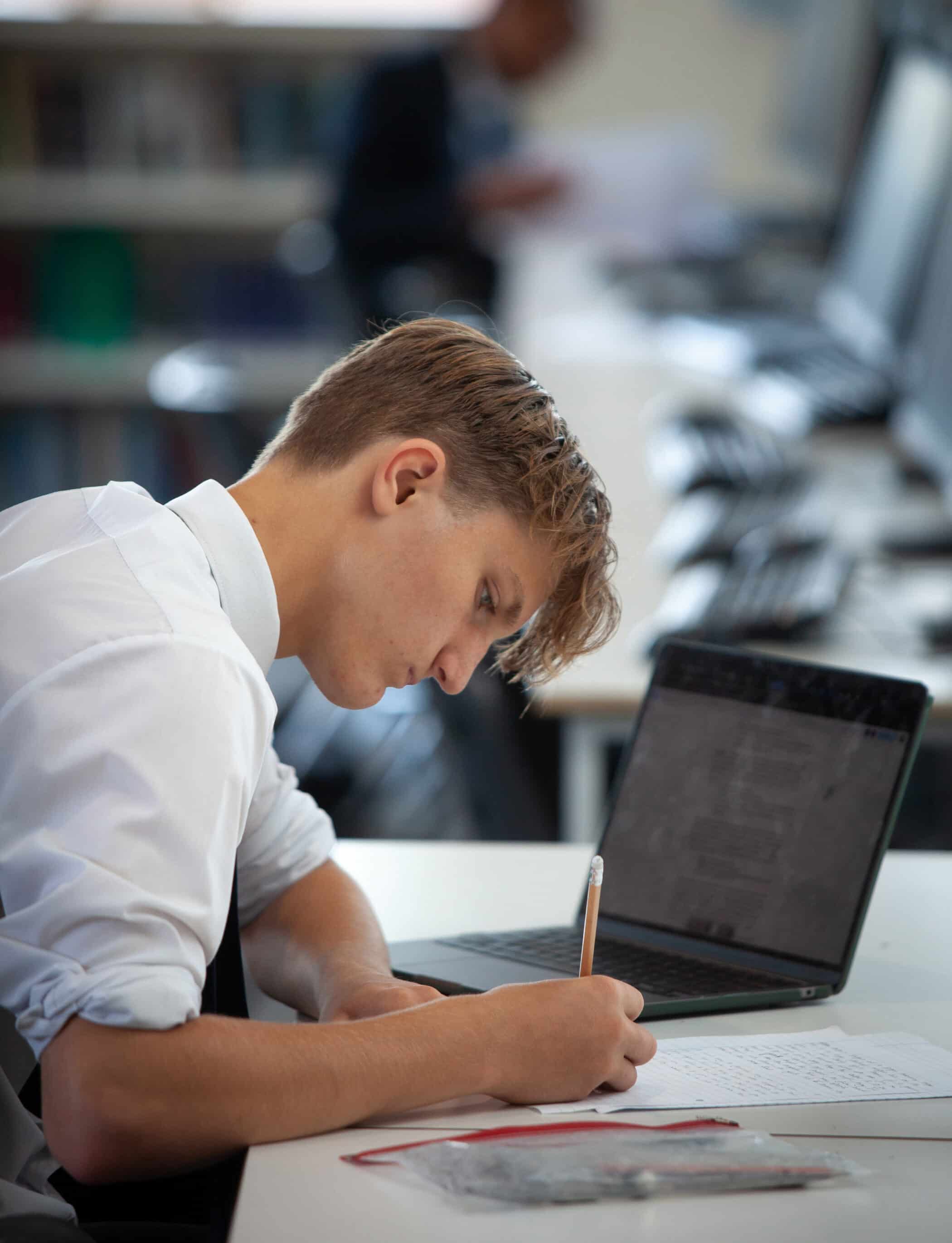 A young man in a white shirt sits at a desk, writing on paper with a pen whilst looking at his laptop screen in the Brentwood School library or study area.