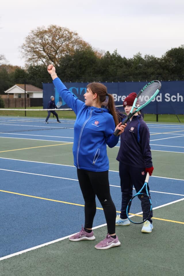 A woman in a blue jacket demonstrates a tennis serve to a child on an outdoor tennis court. The child, also in sportswear, holds a racquet and watches her. Other people play tennis in the background.
