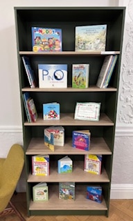 A tall green bookcase displays various children's books facing forwards and stacked upright on five shelves, alongside a tan chair on the left, against a white wall with ornate detailing.