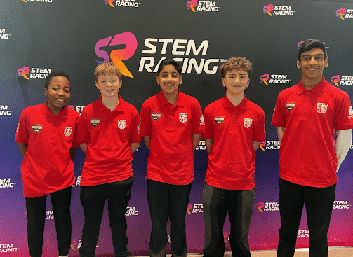 Five smiling students in matching red shirts stand in front of a backdrop with the STEM Racing logo. They are posing for a group photo at what appears to be a STEM Racing event or competition.