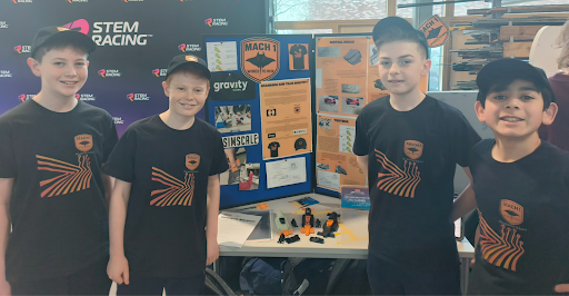 Four boys wearing matching black T-shirts and caps stand in front of a STEM Racing display table. Behind them are project boards, awards, and models, showcasing their team and achievements.