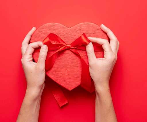 Two hands are tying a red ribbon on a heart-shaped gift box, set against a bright red background.