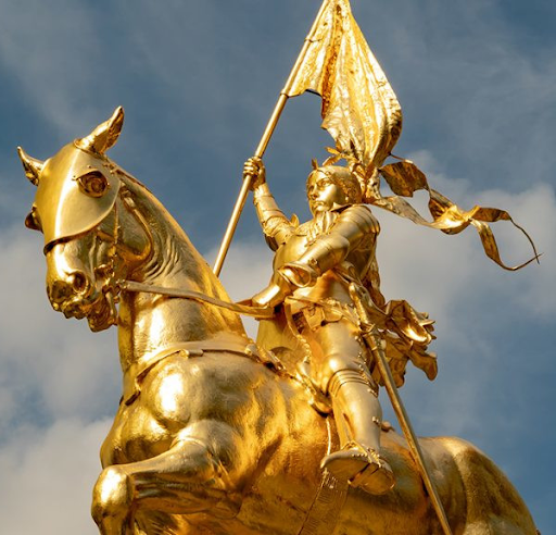 A shiny golden statue of a woman in armour riding a horse and holding a flag, set against a partly cloudy blue sky.
