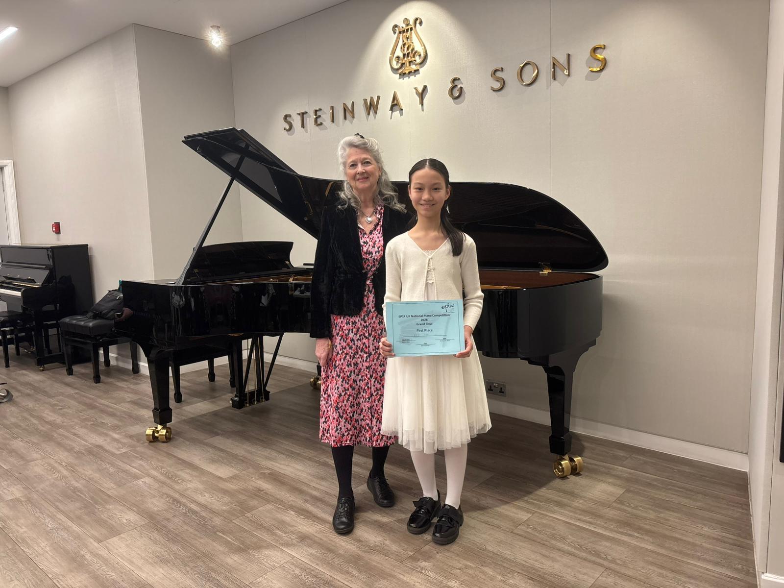 A woman and a girl stand smiling in front of a grand piano at Steinway & Sons; the girl holds a certificate and wears a white dress, while the woman wears a pink-patterned dress with a black jacket.