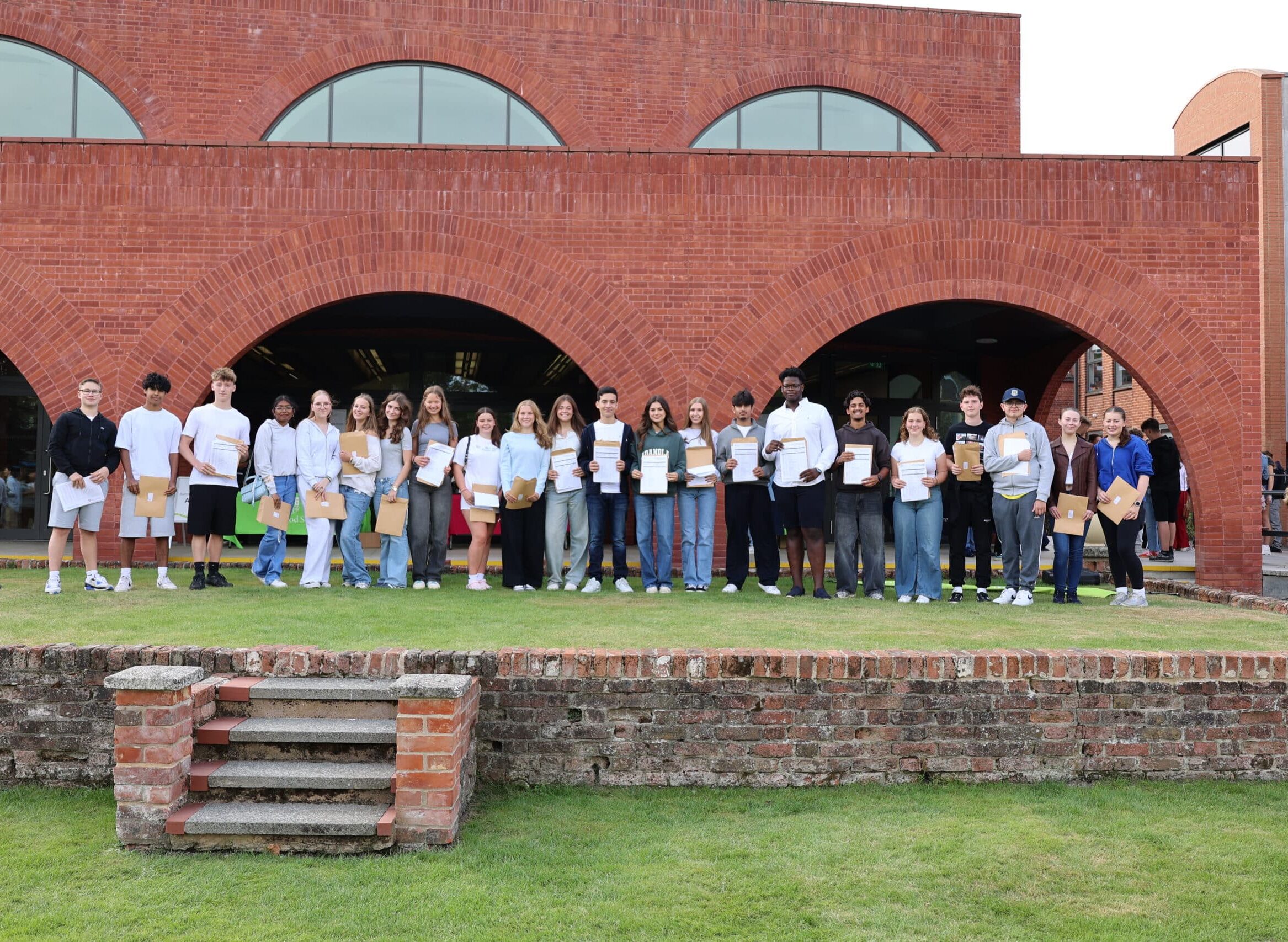 A group of students stand in a row on grass, holding certificates and envelopes, in front of a modern red-brick building with arched windows. Some smile at the camera, celebrating achievements.