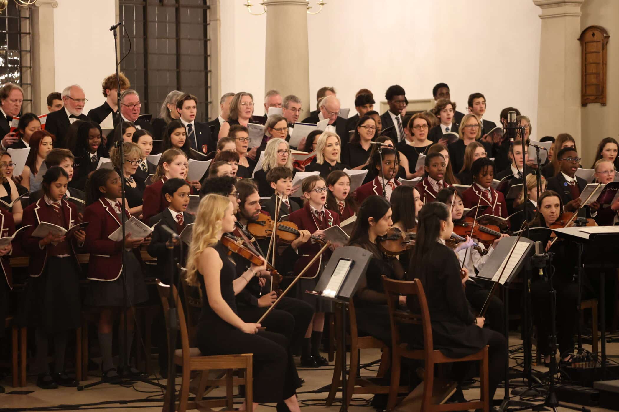 A choir and orchestra perform together in a hall. The choir, featuring adults and children in uniforms, stand behind musicians playing string instruments. Everyone is focussed and engaged in the performance.
