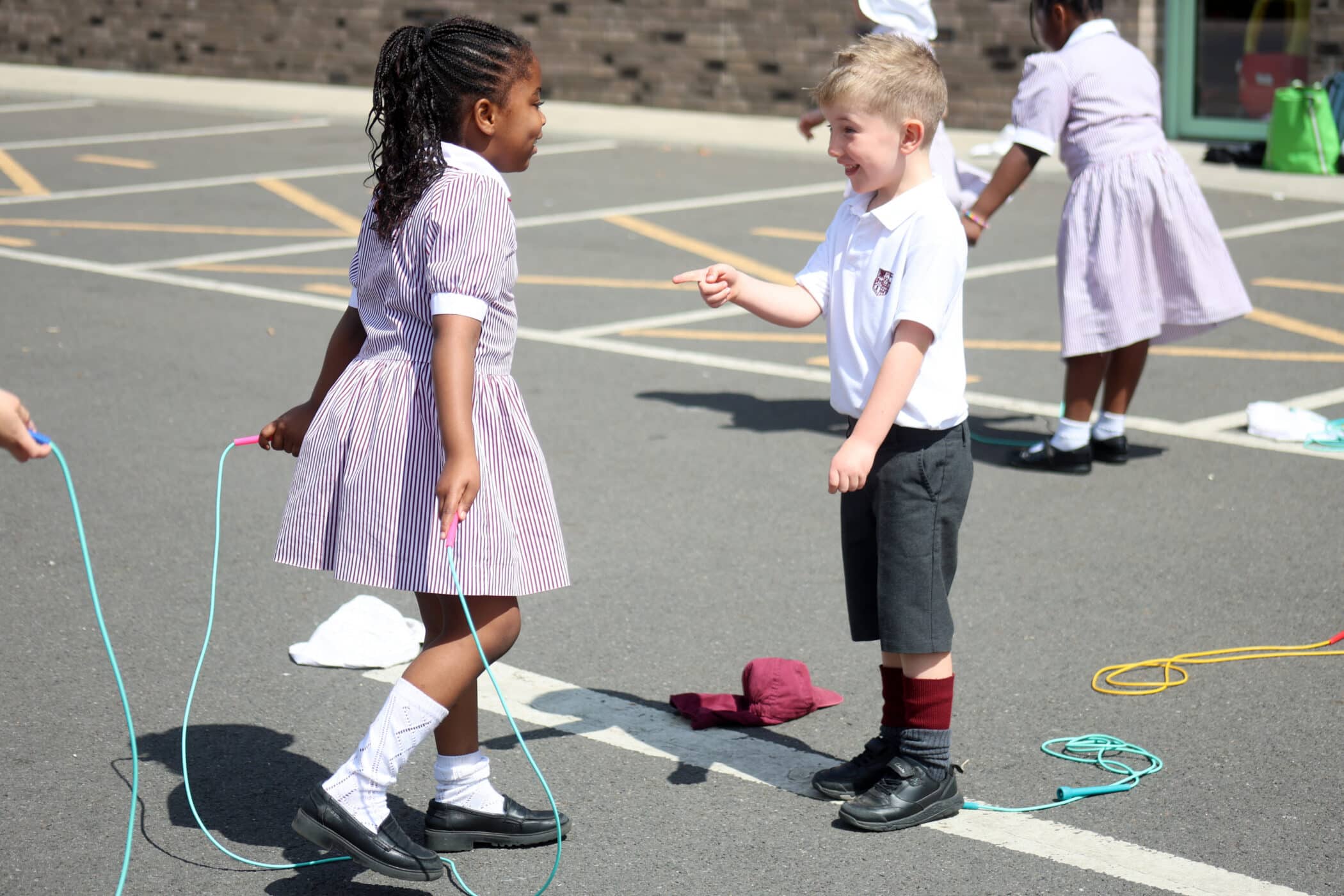 Two young children, one girl and one boy, are smiling and playing with skipping ropes on a sunny day in a school playground. Other children are visible in the background, also holding skipping ropes.