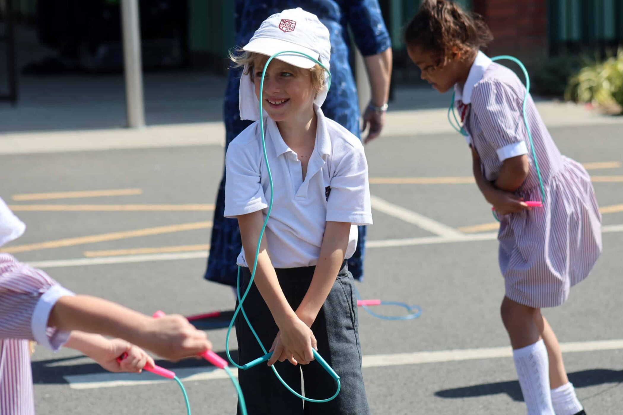 Children in school uniforms play with skipping ropes outdoors on a sunny day. One child in a white hat and polo shirt smiles while skipping, while others participate in the background.