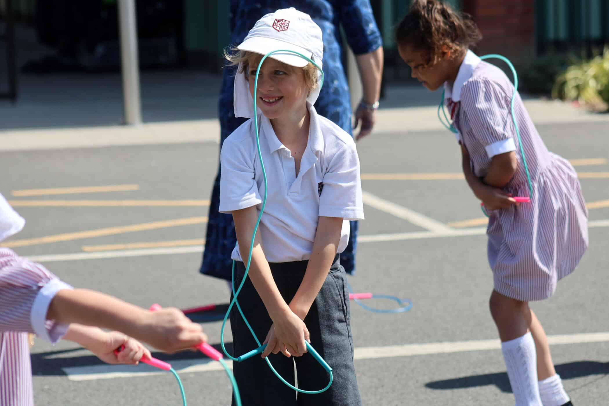 Children in school uniforms smile and play with skipping ropes outside on a sunny day. One child in the centre wears a white cap and holds a rope, whilst others jump nearby.