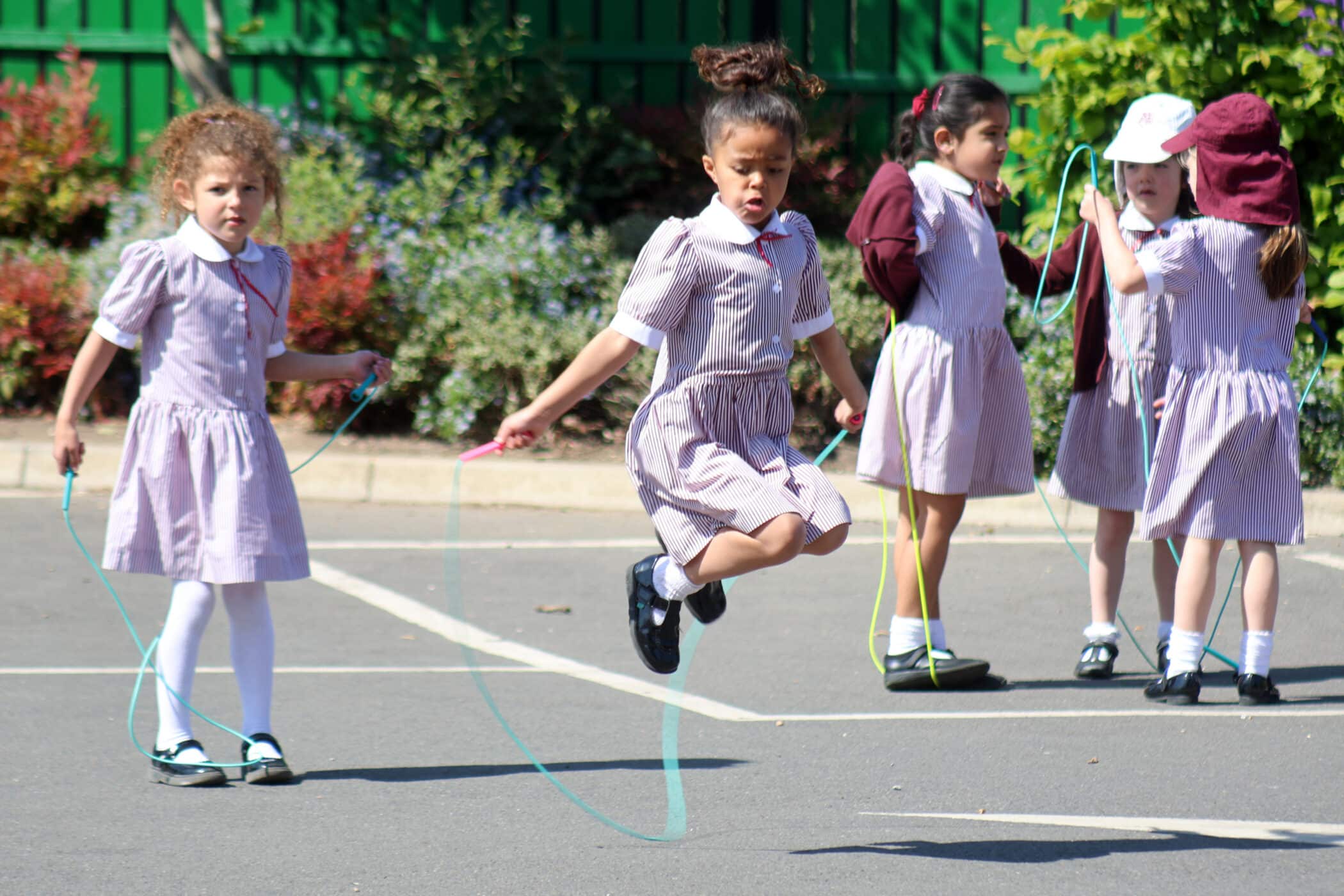 Five young girls in matching school uniforms skip rope and play together outside on a sunny day, with greenery and a fence in the background. One girl is mid-jump, and the others are either skipping or standing nearby.