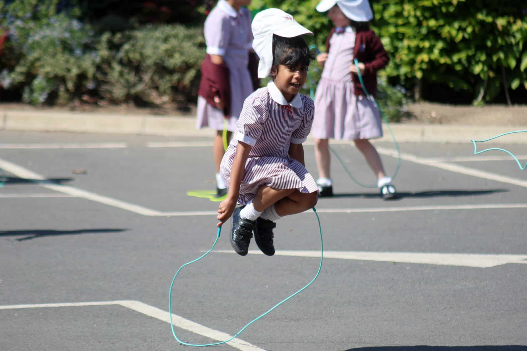 A young girl in a school uniform skips rope and jumps high in the air on a paved playground, with other children in similar uniforms playing in the background.