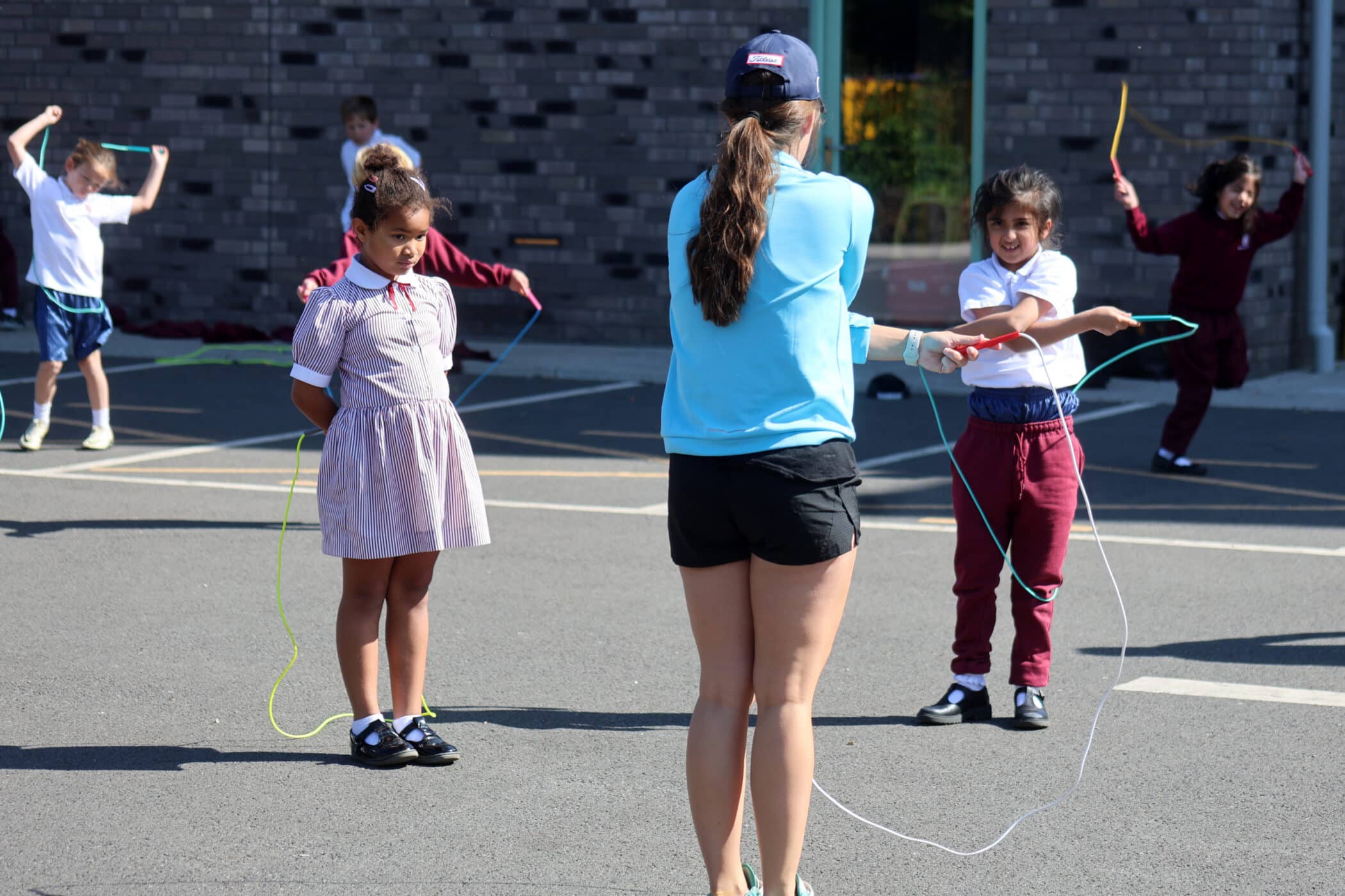 A woman in a blue shirt and shorts instructs a group of children skipping rope outside on a sunny day. Some children are jumping, whilst others await their turn, in a paved area near a building.