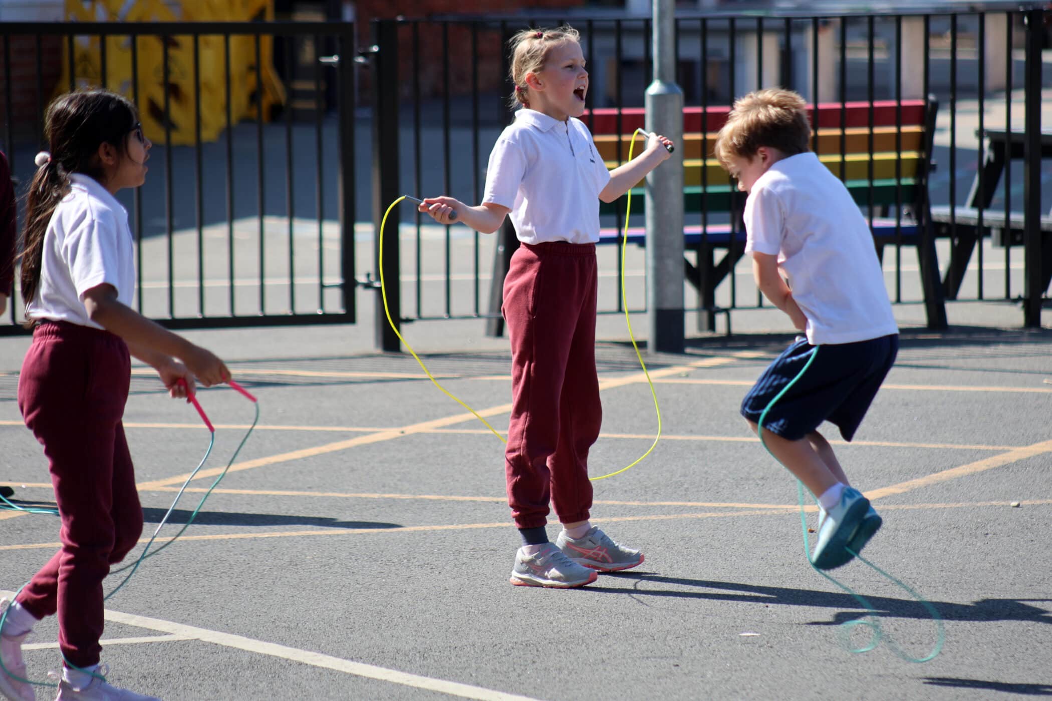 Three children, two girls and one boy, are skipping and playing on a sunny playground. They are wearing school uniforms and appear to be having fun, with one girl smiling and jumping energetically.