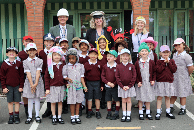 A group of young schoolchildren in uniform and colourful hats stand outside with three adults behind them, also wearing hats. Everyone is smiling and posing for the photo. The background shows a striped building facade.