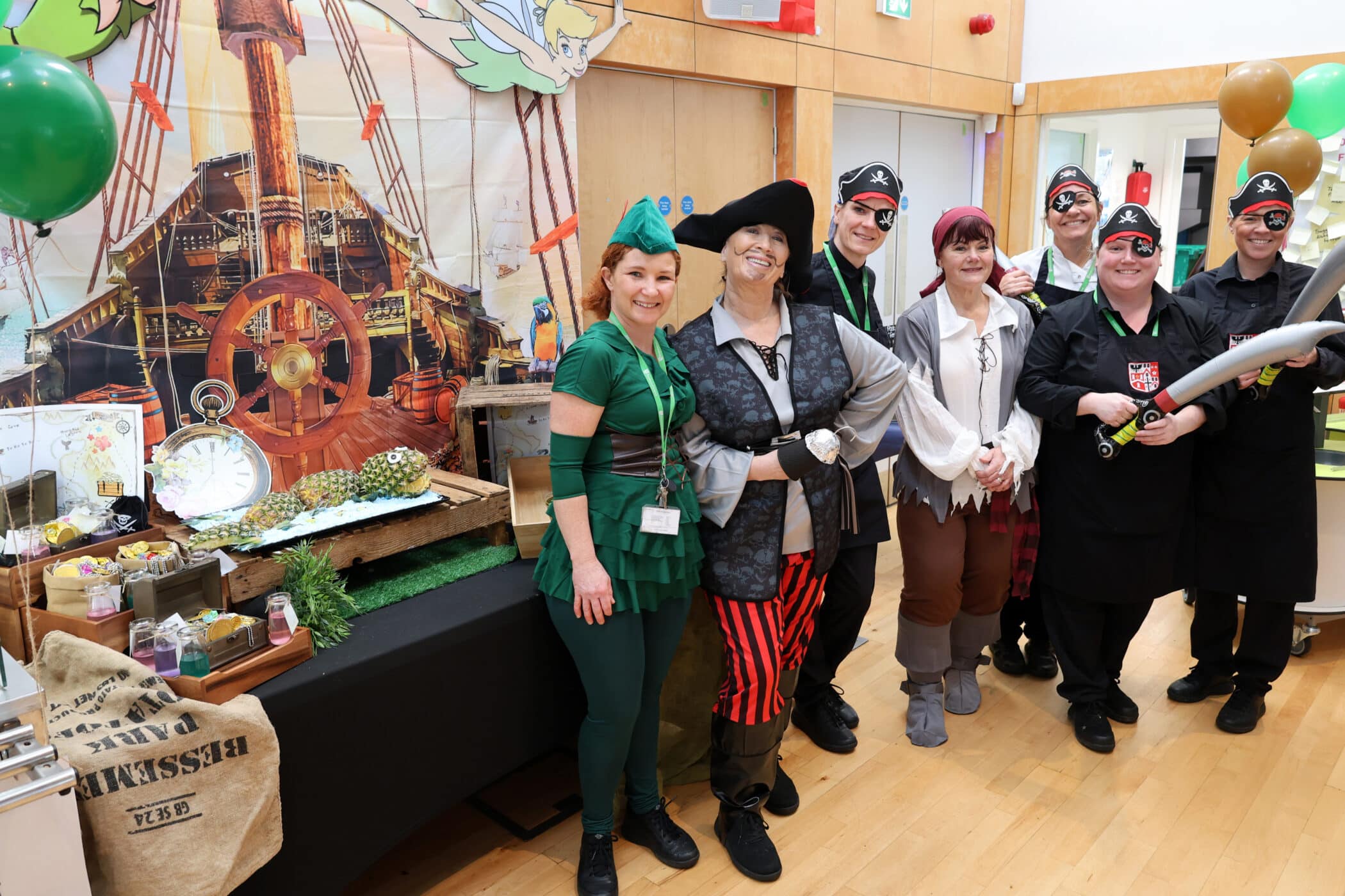 A group of adults dressed as Peter Pan and pirates stand smiling in a decorated room with a pirate ship backdrop, green and brown balloons, and a table with themed food and props.