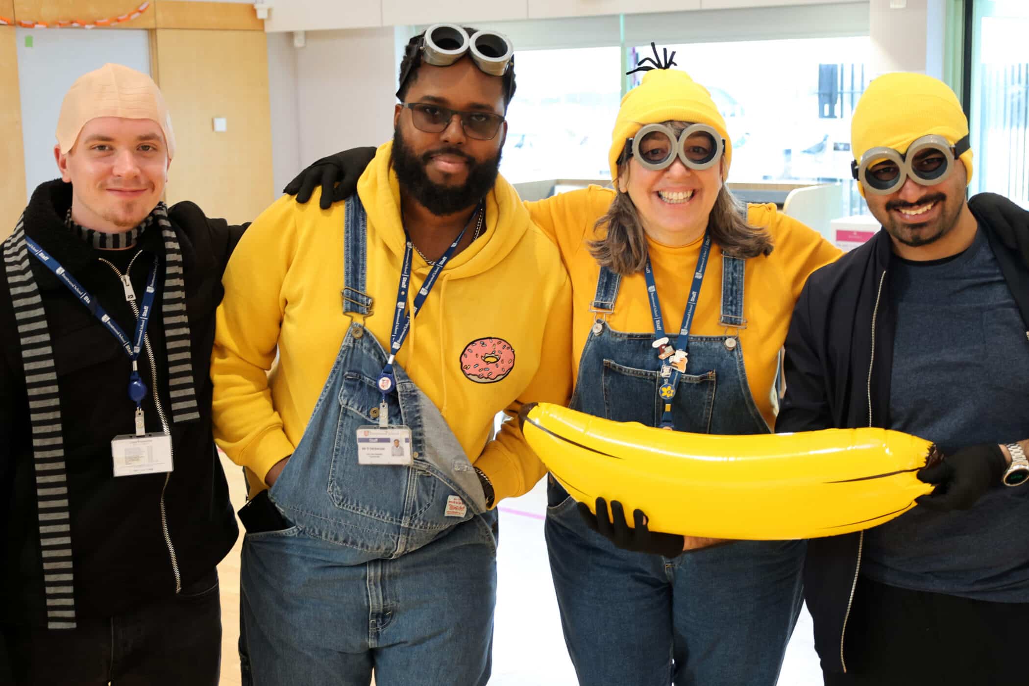 Four adults dressed as Minions from Despicable Me, wearing yellow tops, dungarees, yellow hats or bald caps, and goggles. One holds a large inflatable banana. They are smiling and posing together indoors.