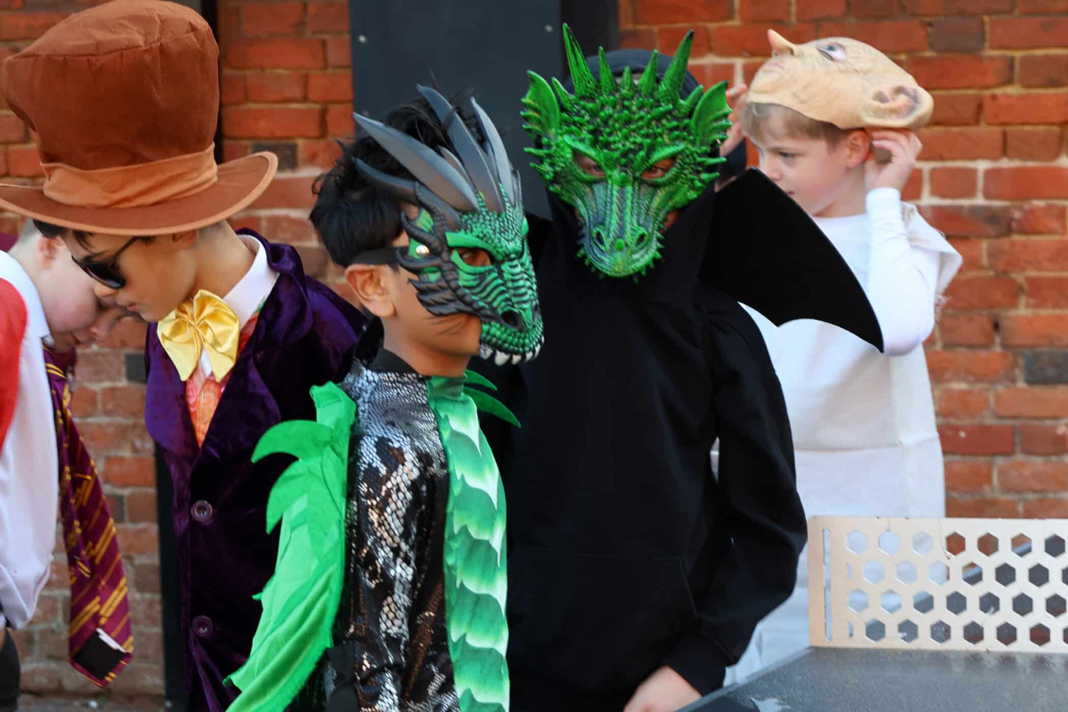 Children in colourful costumes stand together; two wear vibrant green dragon masks and wings, another wears a large brown top hat and purple coat, and a fourth is dressed in white with a pig headpiece. A brick wall is in the background.