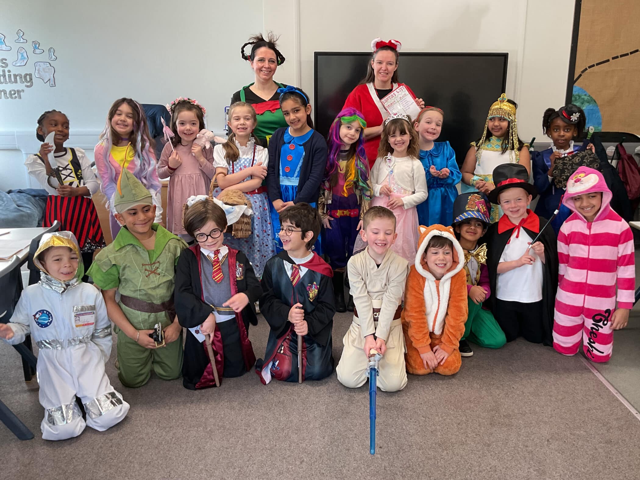 A group of children and two adults in colourful costumes pose and smile for a group photo in a classroom. The children are dressed as various characters, including animals, storybook heroes, and fantasy figures.