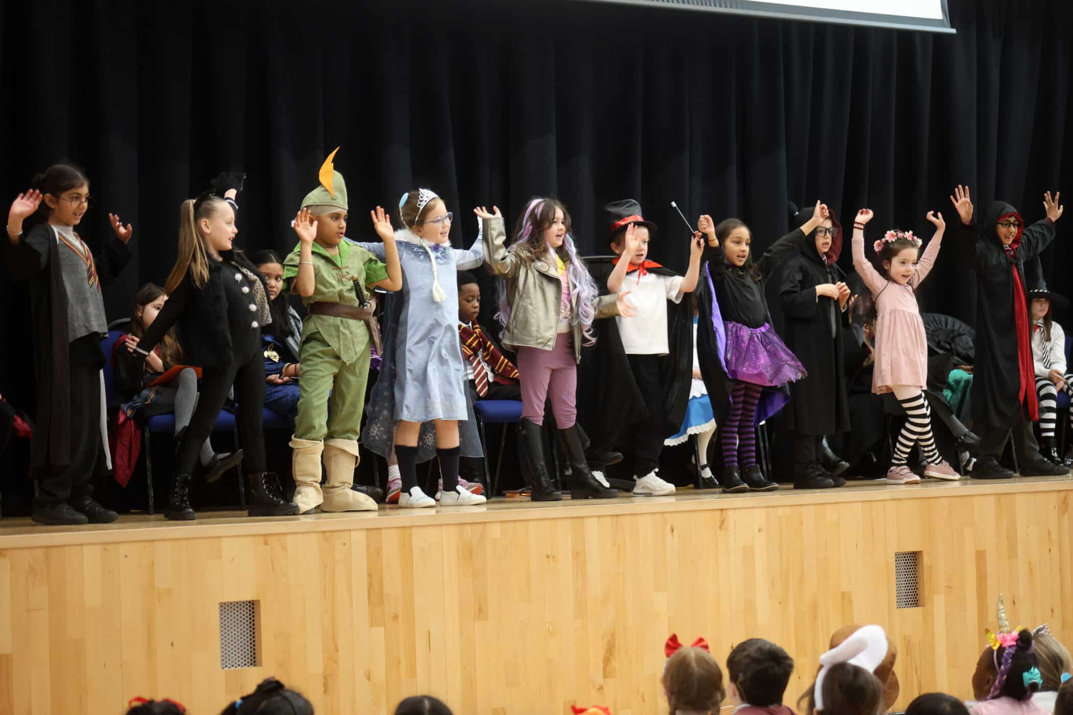 A group of children in colourful costumes stand on a stage, holding hands and raising their arms, performing in front of an audience. The background is a dark curtain, and kids in costume are seated behind them.