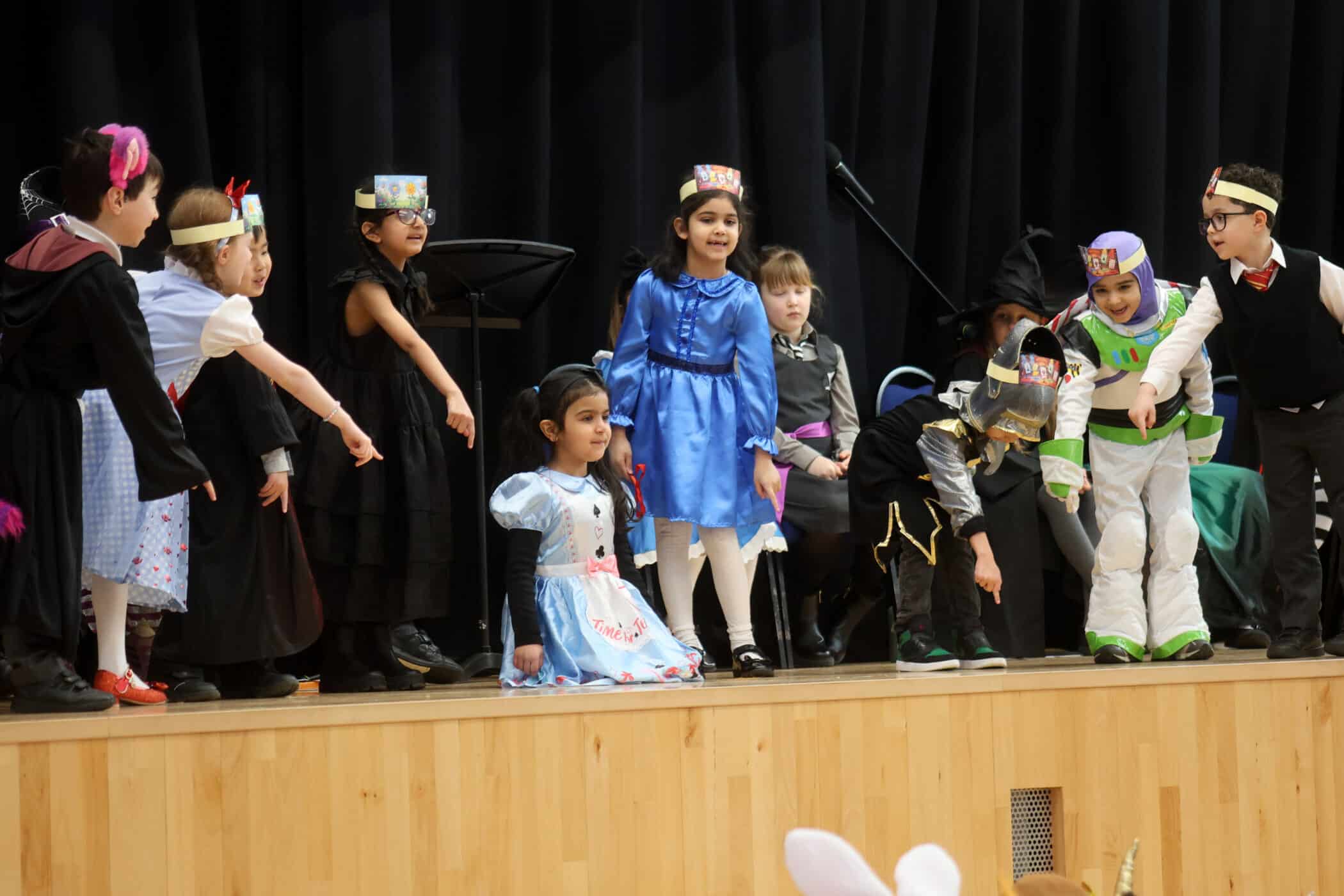 A group of children in colourful costumes stand on a stage, some pointing and some with hands at their sides, against a dark curtain backdrop during a school play or performance.