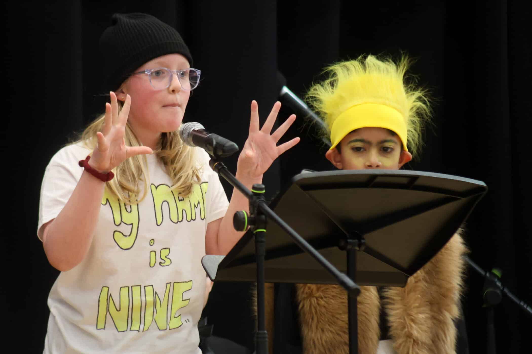 Two children perform on stage. One speaks into a microphone, gesturing with raised hands, wearing glasses, a black beanie, and a shirt reading My name is NINE. The other wears a yellow wig and furry costume, standing by a music stand.