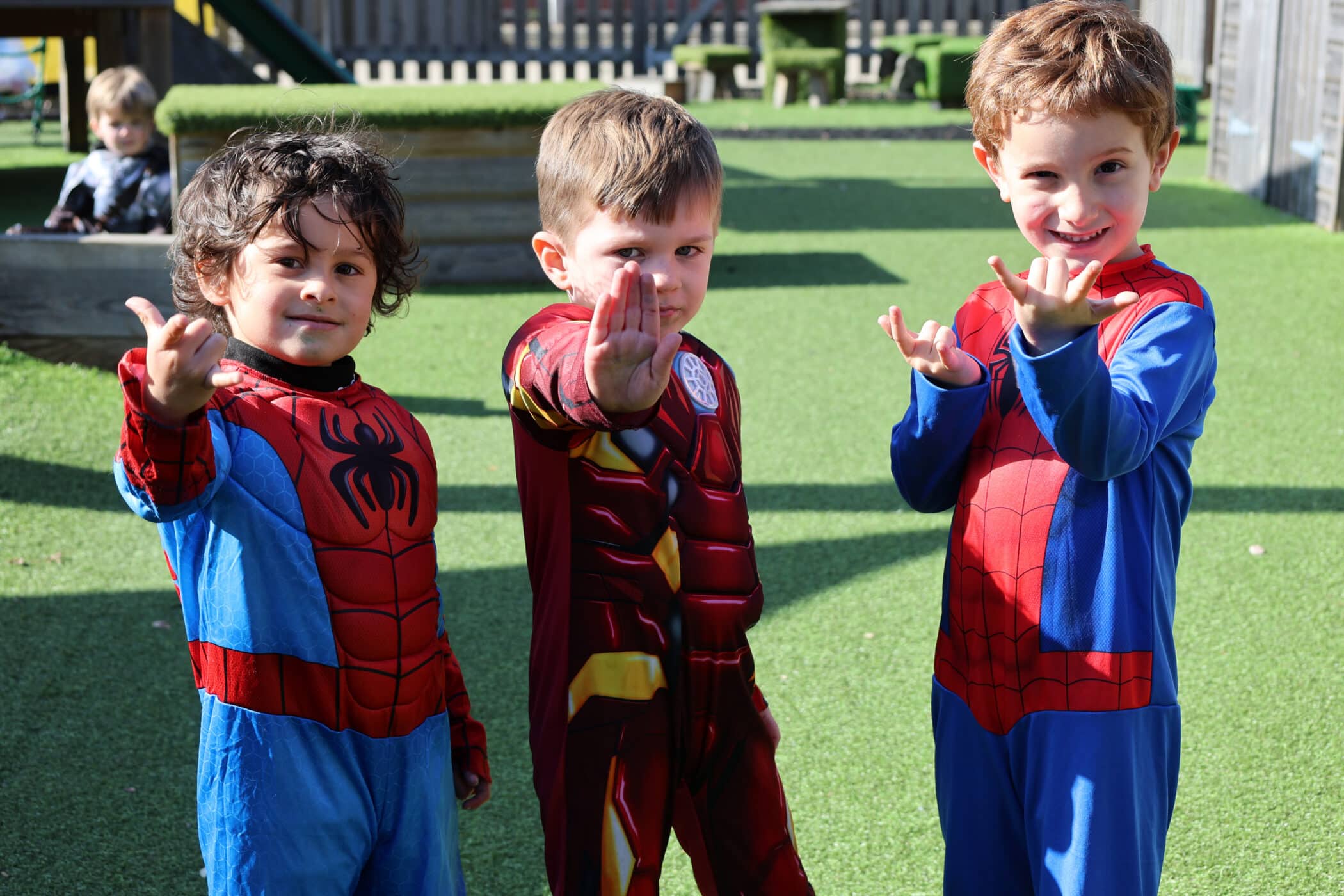 Three young children in superhero costumes, two as Spider-Man and one as Iron Man, stand outside on green artificial grass, making playful superhero poses and smiling at the camera. A child sits in the background.