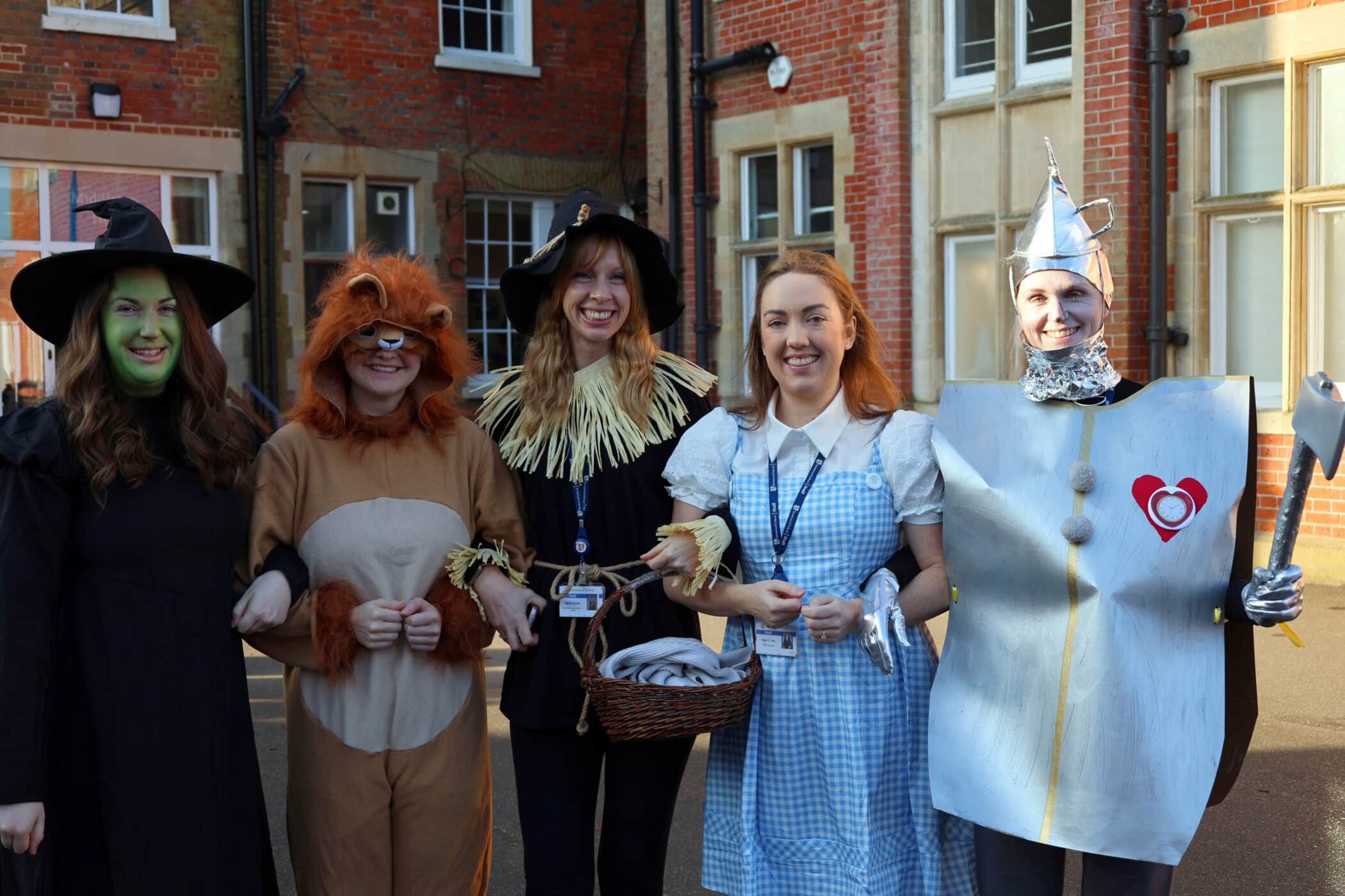 Five people dressed as characters from The Wizard of Oz—the Wicked Witch, Cowardly Lion, Scarecrow, Dorothy, and Tin Man—stand smiling and arm in arm outside a brick building.