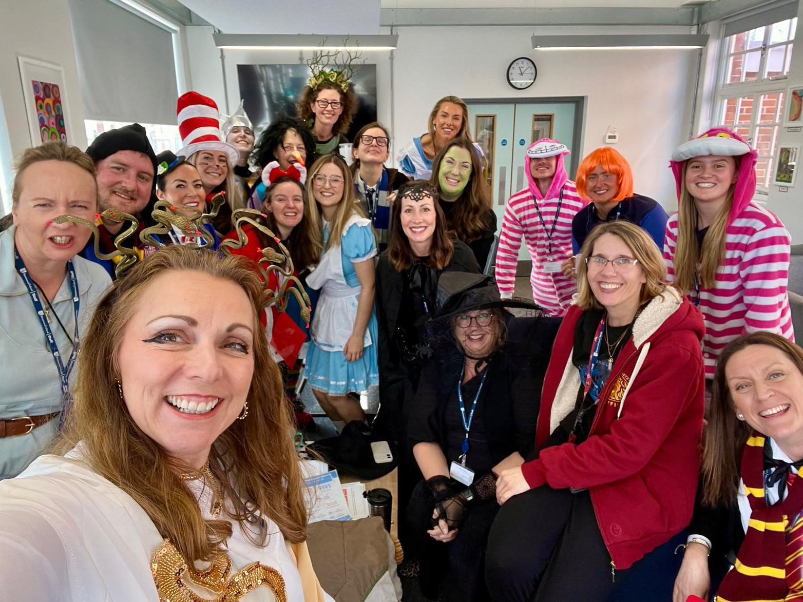 A group of adults dressed in colourful costumes, including storybook characters and witches, pose together indoors, smiling for a selfie in a bright classroom with large windows and a clock on the wall.