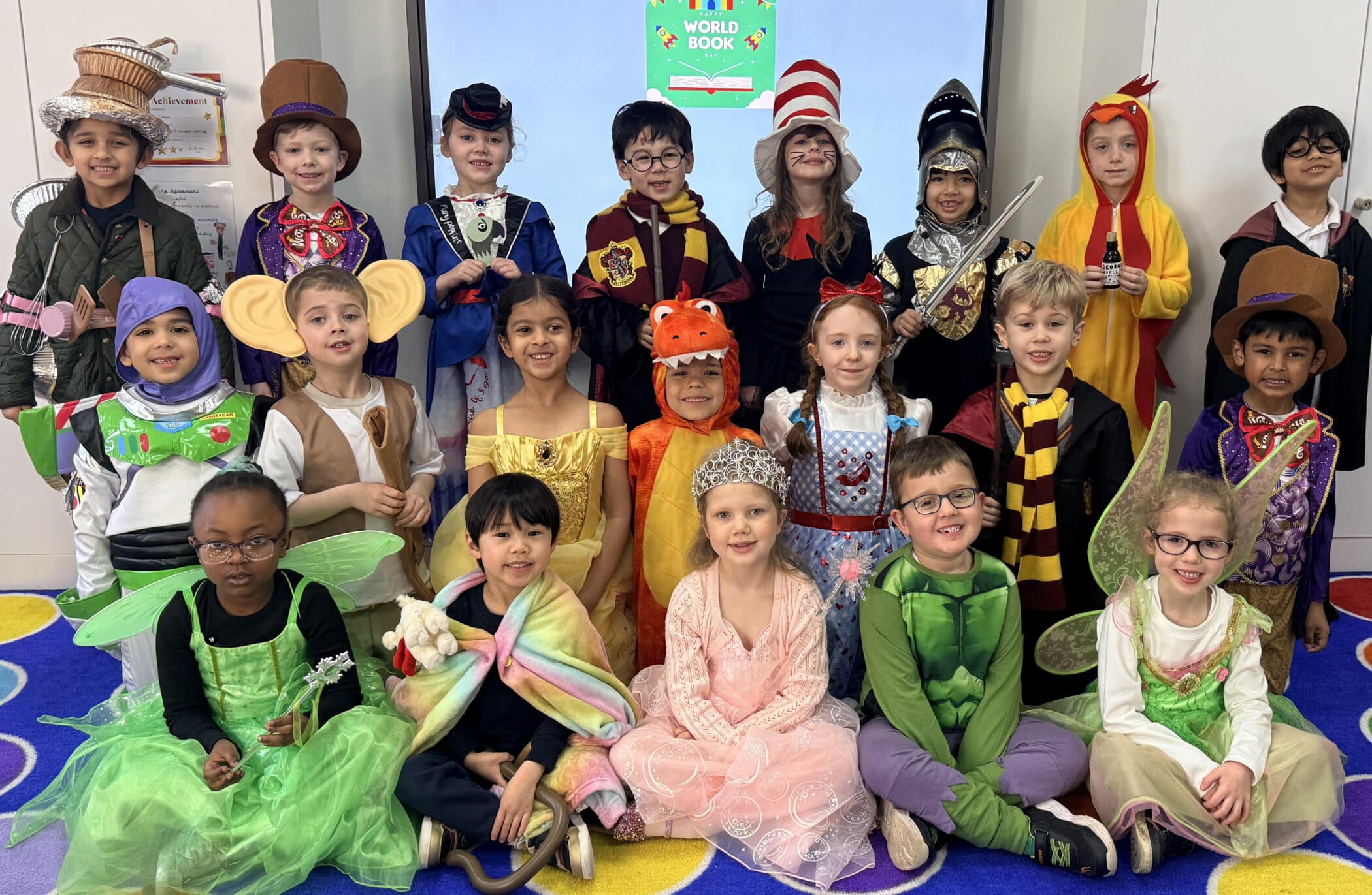 A group of children dressed in colourful costumes, including fairies, princesses, superheroes, and book characters, smile and pose together in a classroom with a World Book display in the background.