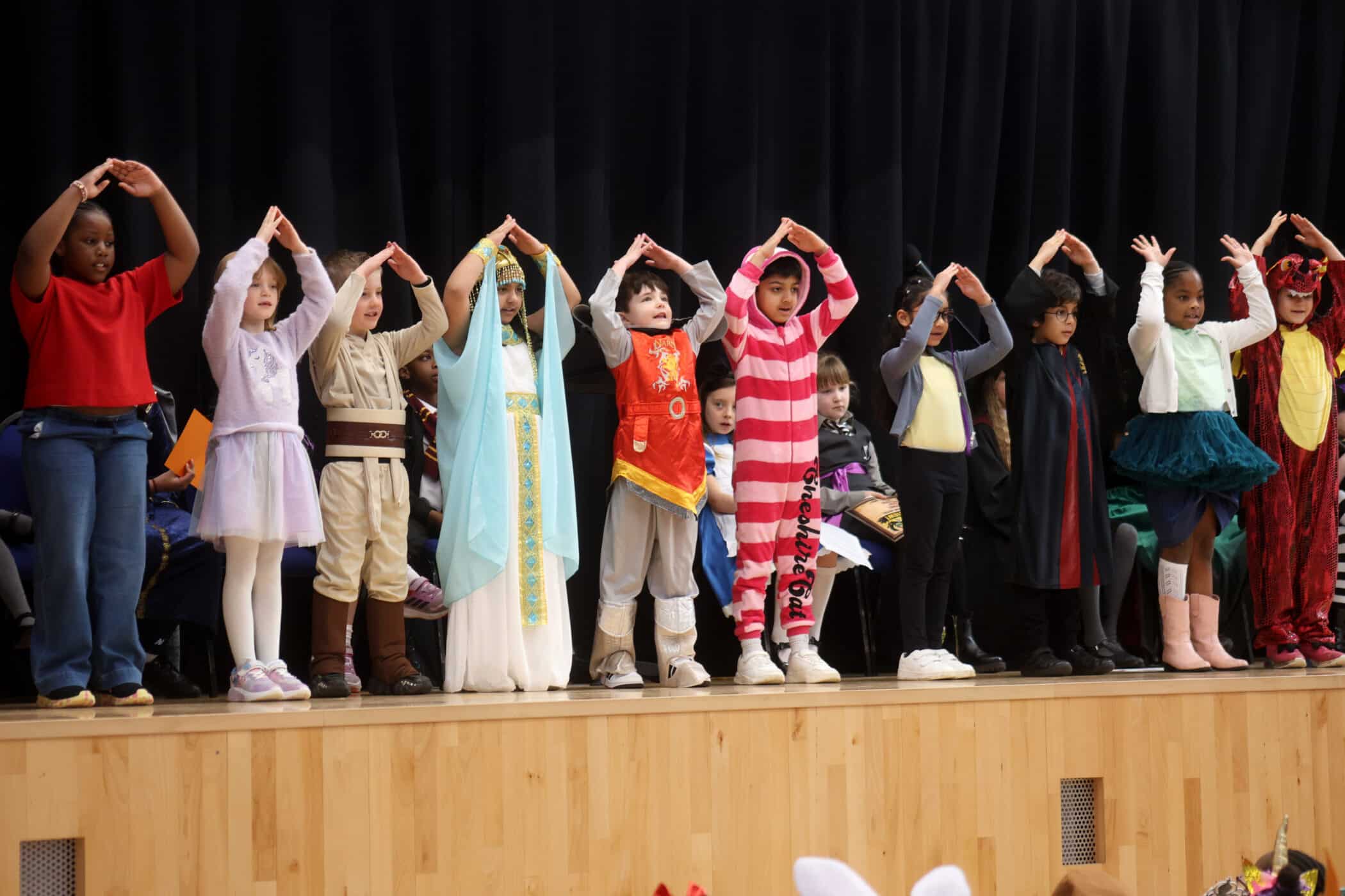 A group of children in colourful costumes stand on a stage with their arms raised above their heads, forming triangles. A black curtain is in the background.